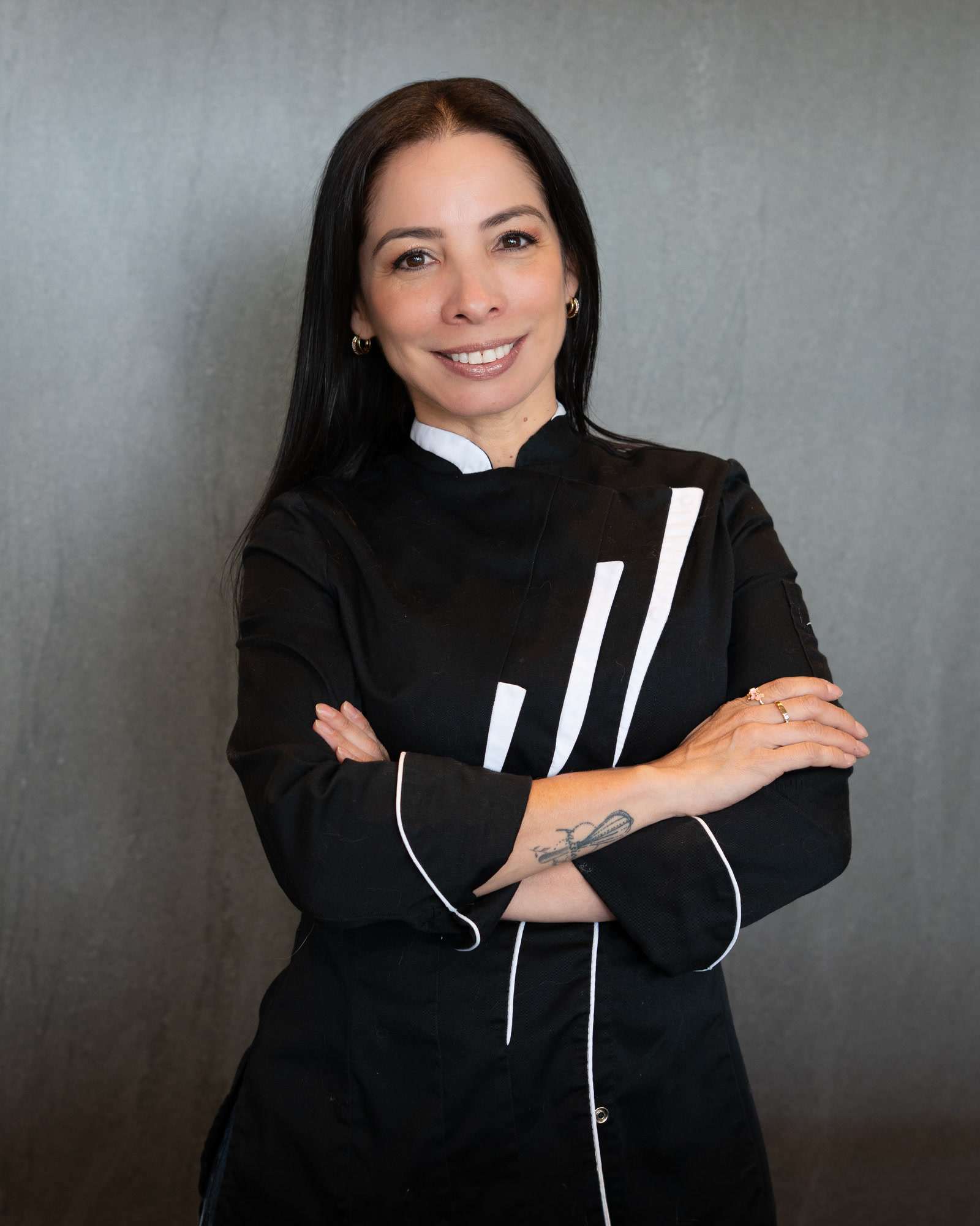 Juliana Romero, Chef July, con chaqueta de chef negra y los brazos cruzados, sonríe frente a un fondo gris.