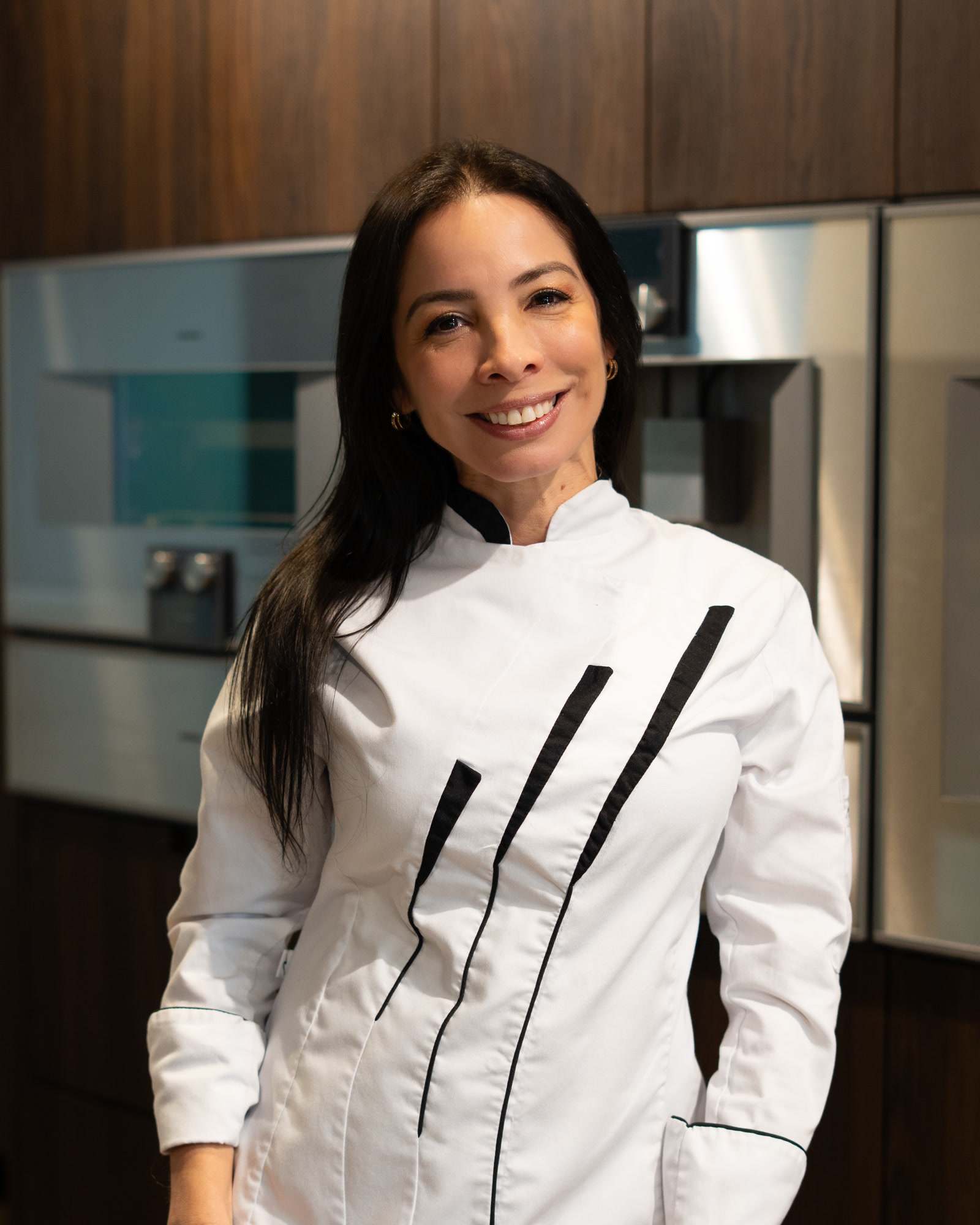 Juliana Romero sonríe con chaqueta de chef blanca de líneas negras, frente a muebles de madera y hornos de acero.