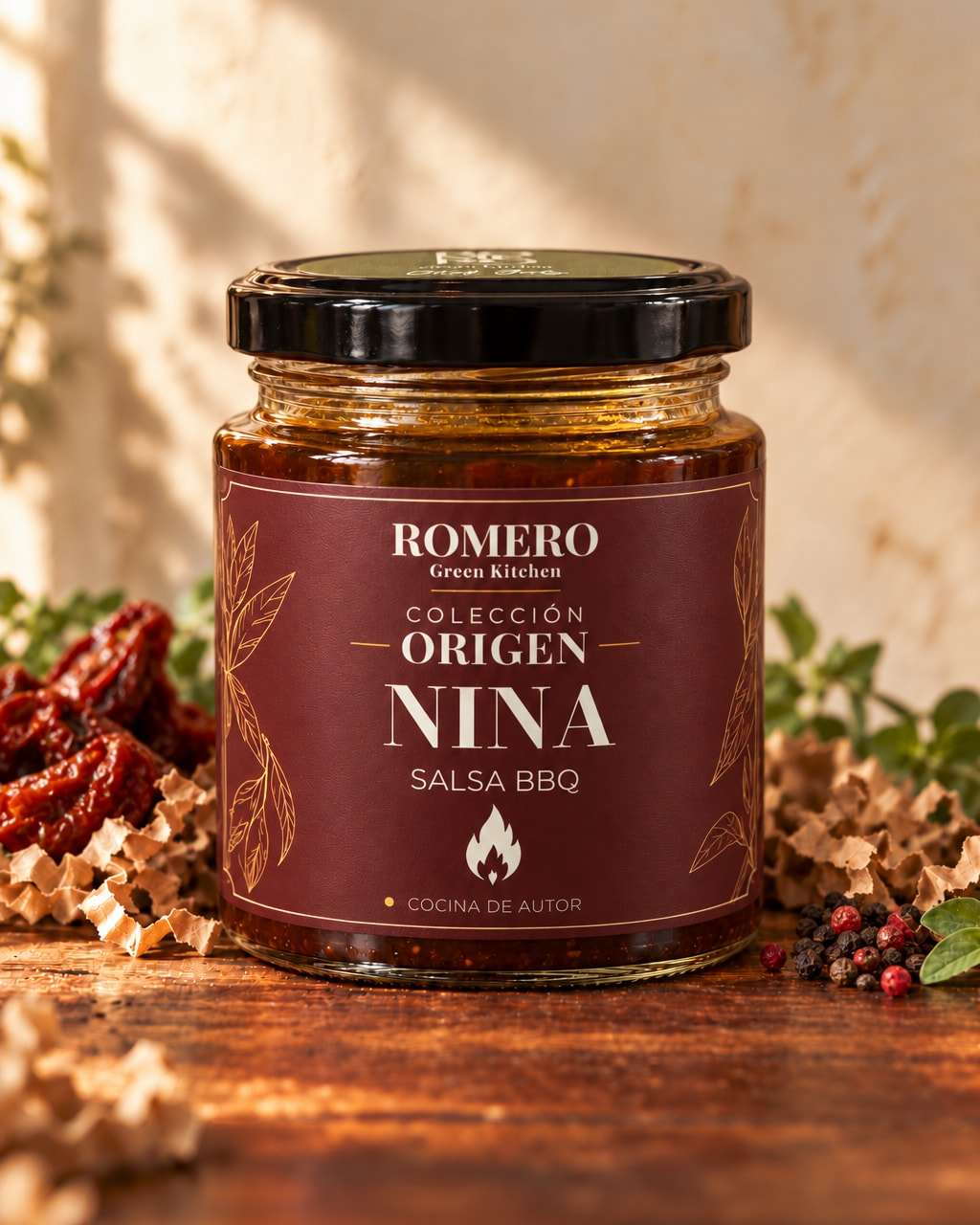Frasco de Nina, salsa BBQ oscura, con etiqueta burdeos de Romero Colección Origen y tapa negra, junto a tomates secos y pimienta en grano.